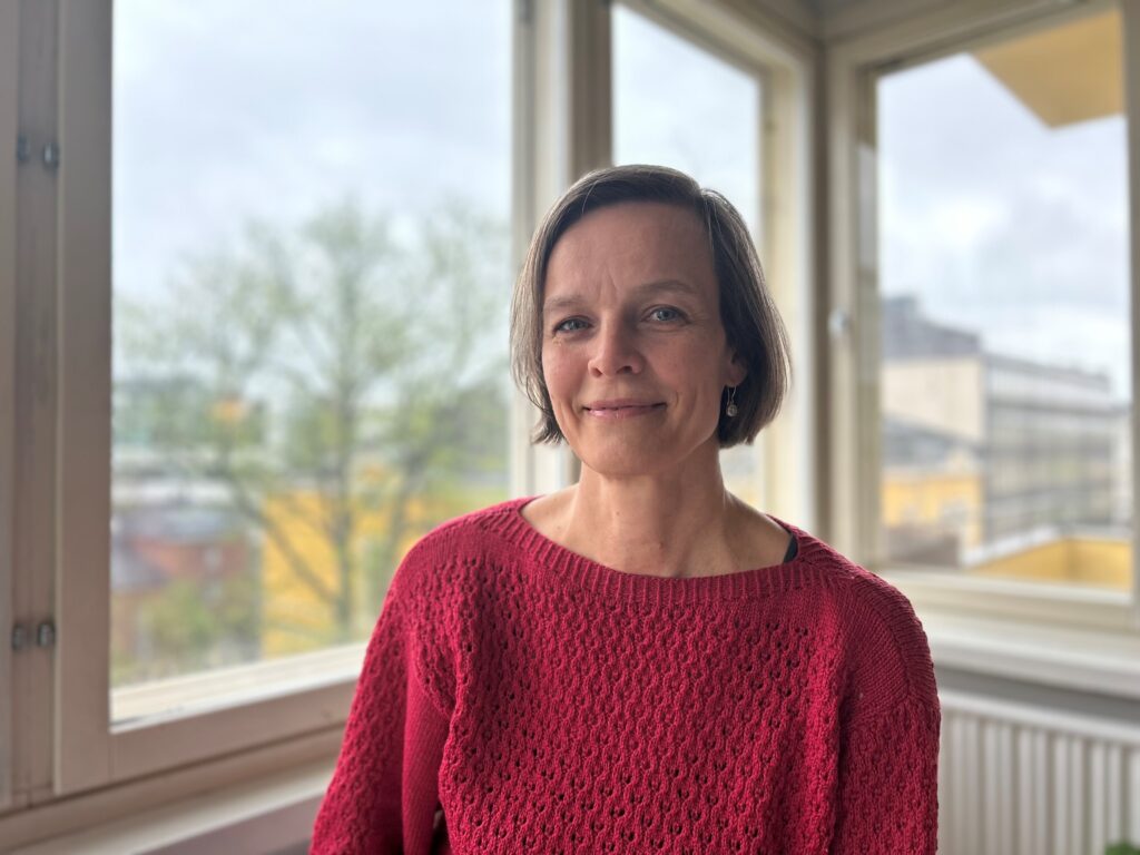 Bild på Anna-Lena Sahlström som står framför ett fönster (kvinna med brun pagefrisyr iklädd röd tröja).