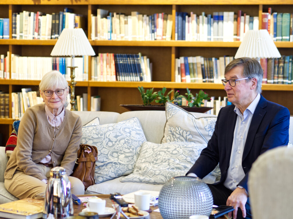 Kvinna med vitt hår, beige tröja och glasögon sitter i soffa tillsammans med en man med gråsprängt hår, mörk kavajrock och ljusblå skjorta. De sitter i ett bibliotek, med en Kanne kaffe framför sig på bordet.
