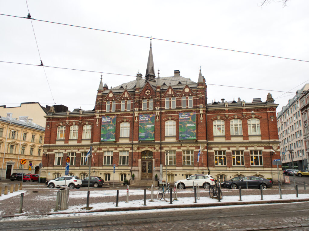 Hus i rött tegel som finns på Högbergsgatan i Helsingfors. Framför huset syns bilar, lite snö på marken, samt en del av gatan. 