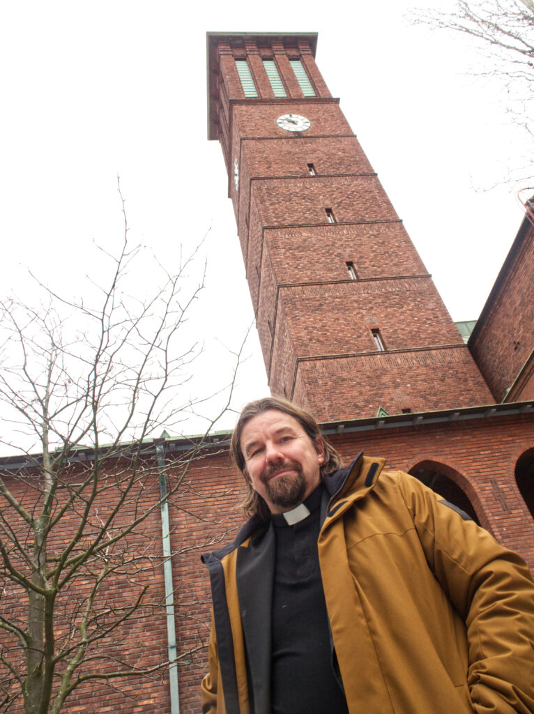 Bild av manlig präst iklädd prästkrage och senapsgul vinterjacka, stående framför en kyrka i rött tegel. Kyrktornet syns bakom honom.