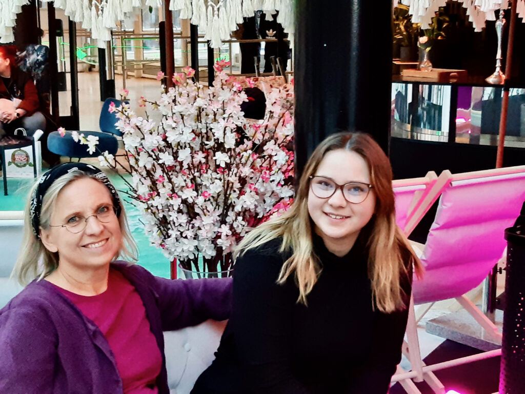 En äldre kvinna (mamma) med glasögon och en yngre kvinna (dottern) tittar in i kameran och ler.