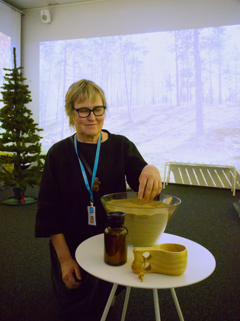 Blond korthårig kvinna står med handen och rör vid sand som finns i en glasskål. Bakom henne finns en plastgran och en vägg där en bild av ett landskap från Lappland projiceras.