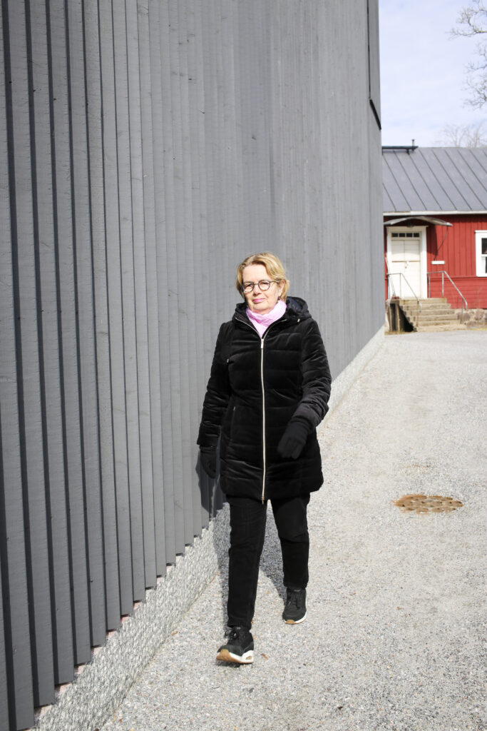 Kvinna i svart rock går på en gata invid en grå trävägg, i bakgrunden skymtar ett rött trähus.