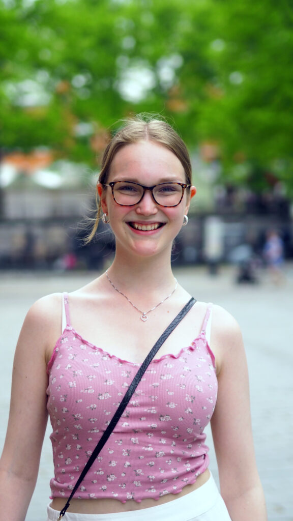 Ung leende kvinna med glasögon och örhängen står utomhus med ett träd bakom sig.