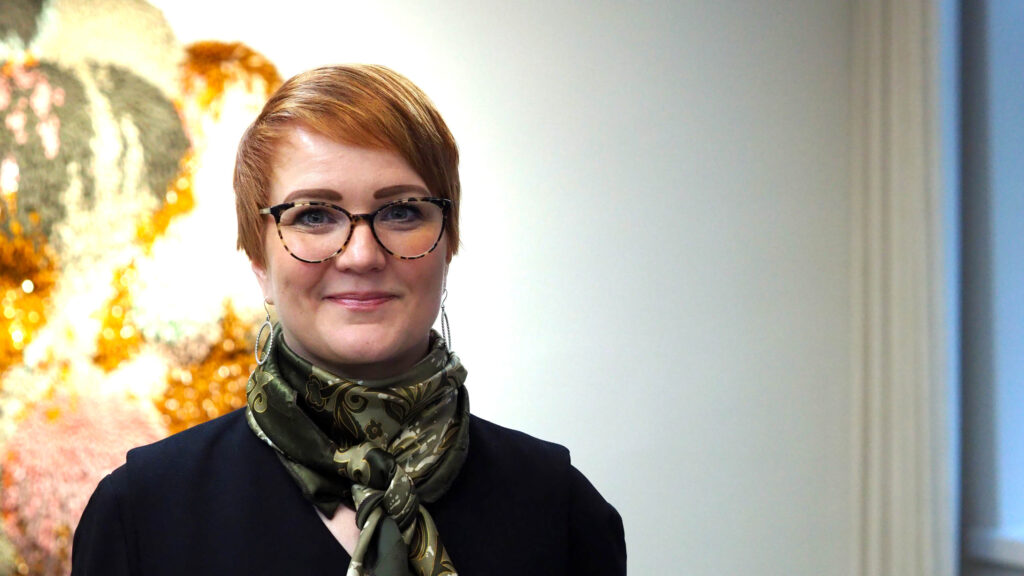På bilden syns en kvinna med kort rött hår och glasögon. Hon har glasögon och en grön scarf och en mörkblå blus. Bakgrunden är vit till höger och suddigt mångfärgad till vänster.