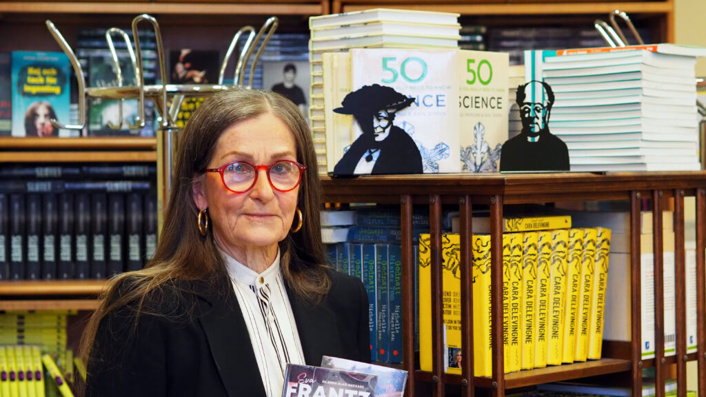 Glasögonprydde kvinna med långt hår står i ett skolbibliotek och håller en bok i handen.