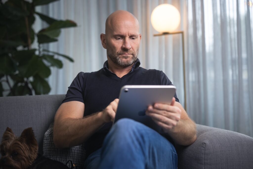 På bilden syns en skallig man i medelåldern som sitter och tittar på en pekplatta. Han har svart kortärmad tröja och jeans. I bakgrunden syns en grå gardin framför ett fönster, en grön krukväxt och en lampa i form av en boll.