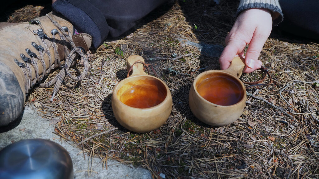På bilden syns två kuksor, det vill säga muggar täljda av trä, som är fyllda med brunt te. De står på ett berg med en matta av bruna granbarr. Man ser en hand som håller i den ena muggens öra.