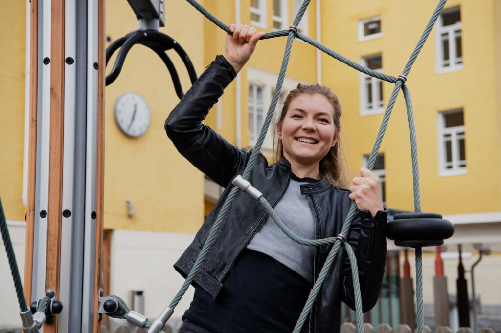 På bilden syns en glad kvinna som står och håller i rep från en klätterställning. Hon har brunt mellanlångt hår och är klädd i en kort svart läderjacka, en grå tröja och svarta byxor. I bakgrunden syns en gul skolbyggnad.