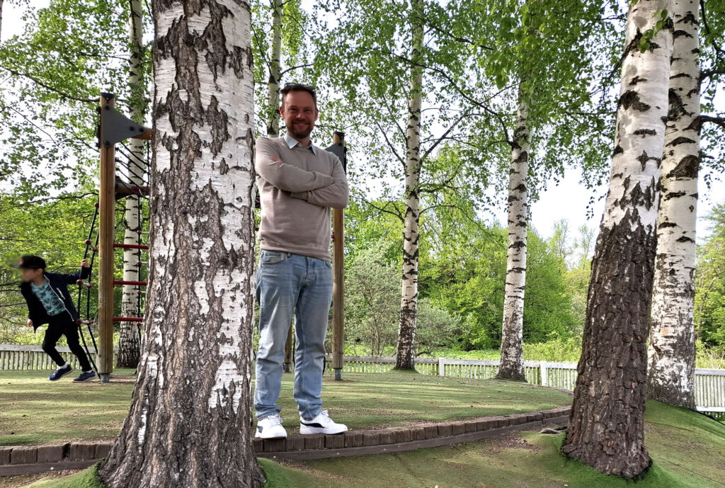 På bilden syns en man som står med händerna i kors. Han har kort skägg och brunt kort hår. Han är klädd i beige tröja, ljusblå jeans och vita sneakers. Runtomkring honom finns stora bjärkstammar och kvistar med gröna blad. I bakgrunden finns en lekställning, och man ser en liten pojke som springer.