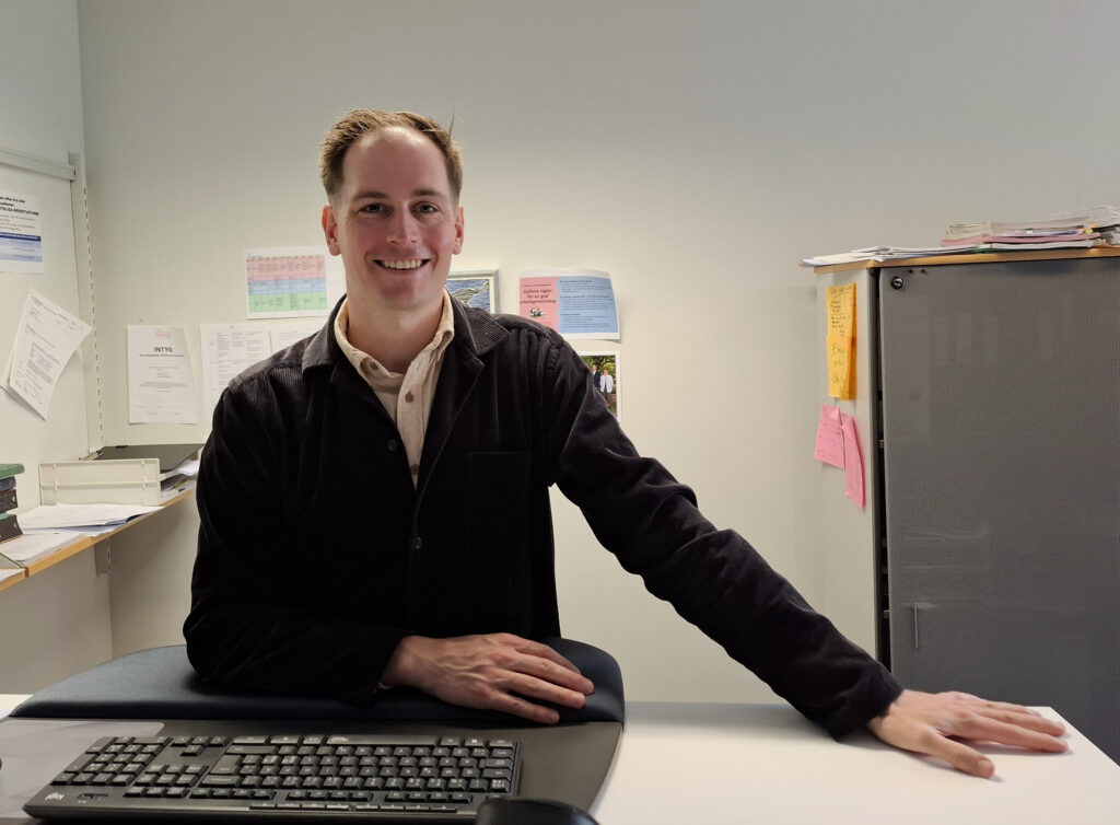 På bilden syns en rätt ung man med hår som börjar på halva flinten. Han ser glad ut. Han har ljus skjortblus och en märk tröja över den. Han sitter vid ett bord och lutar handen på en tjock musmatta. Framför finns ett tangentbord. Bakgrunden är en ljus vägg, där det finns olika papper uppsatta. Man ser också ett skåp med färggranna postits på.