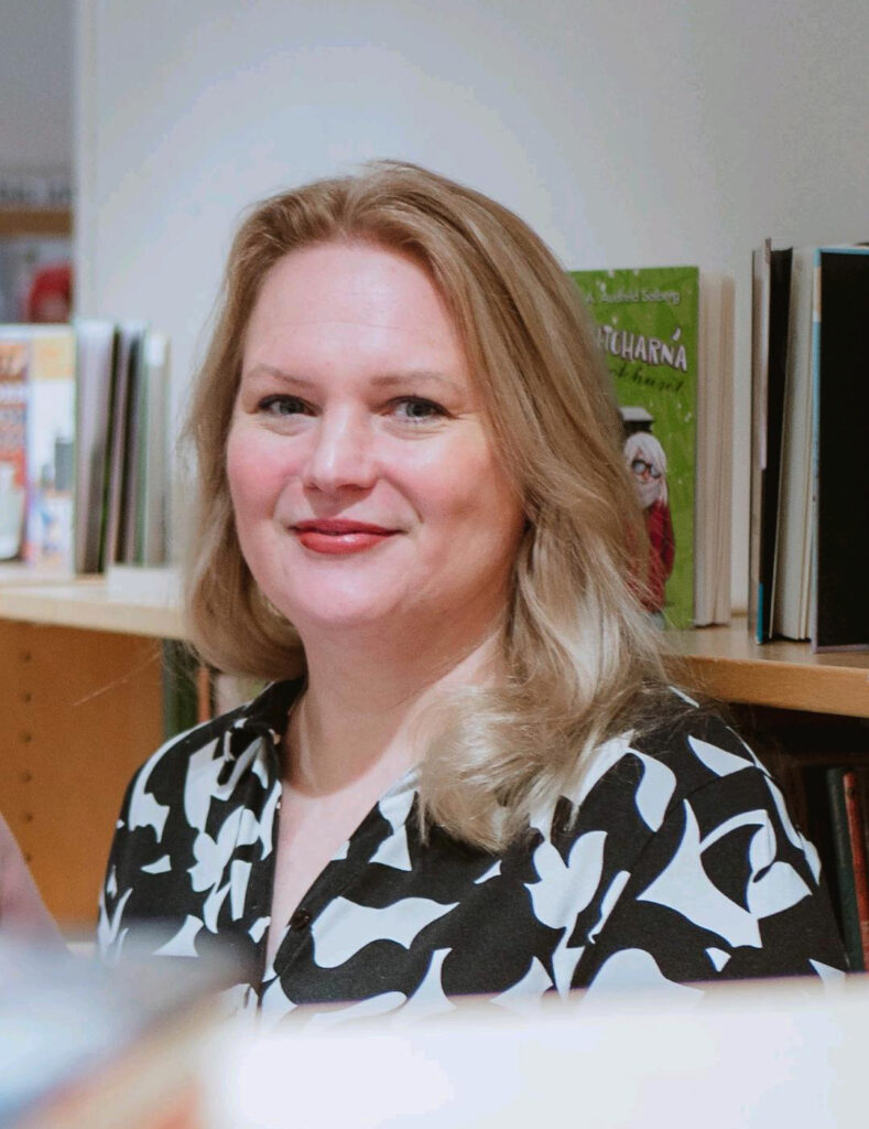 På bilden syns en kvinna som ser glad ut. Hon är fotograferad framför en bokhylla. Hon har en svartvit blus och blont hår.