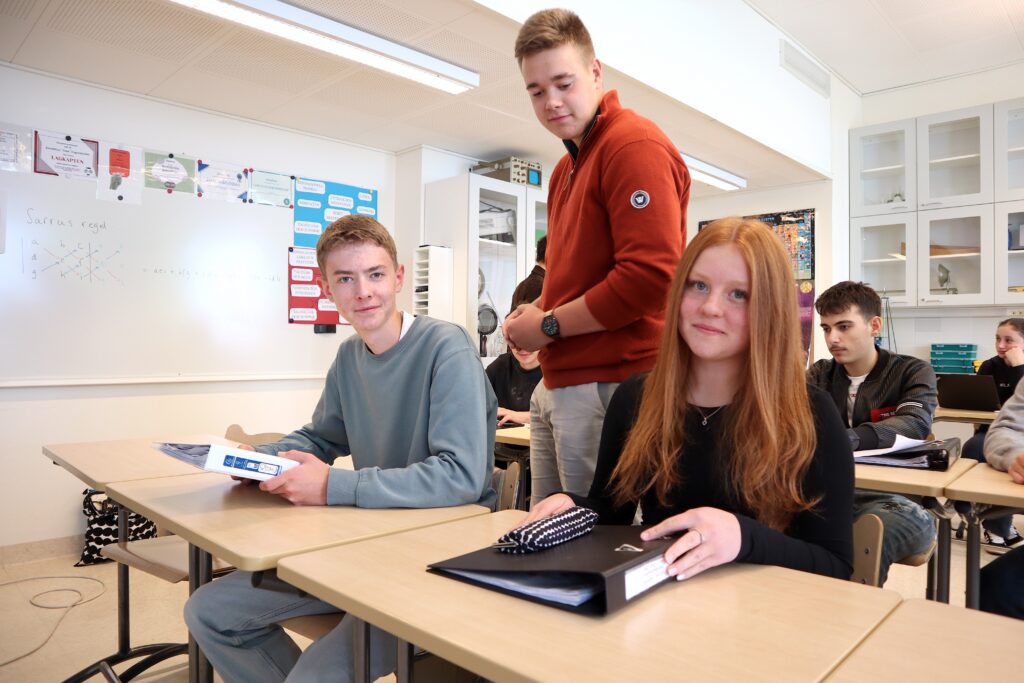 På bilden syns ett klassrum med vita väggar. ljusbrund golv och pulpeter i ljust trä. Till vänster syns en ung man i ljust grönblå collegetröja och jeans och till höger om honom sitter en flicka med långt rött hår och en svart blus. De tittar in i kameran och ler. Bakom dem står en pojke i röd tröja och beigea chinos. Han tittar ner mot de andra två eleverna.