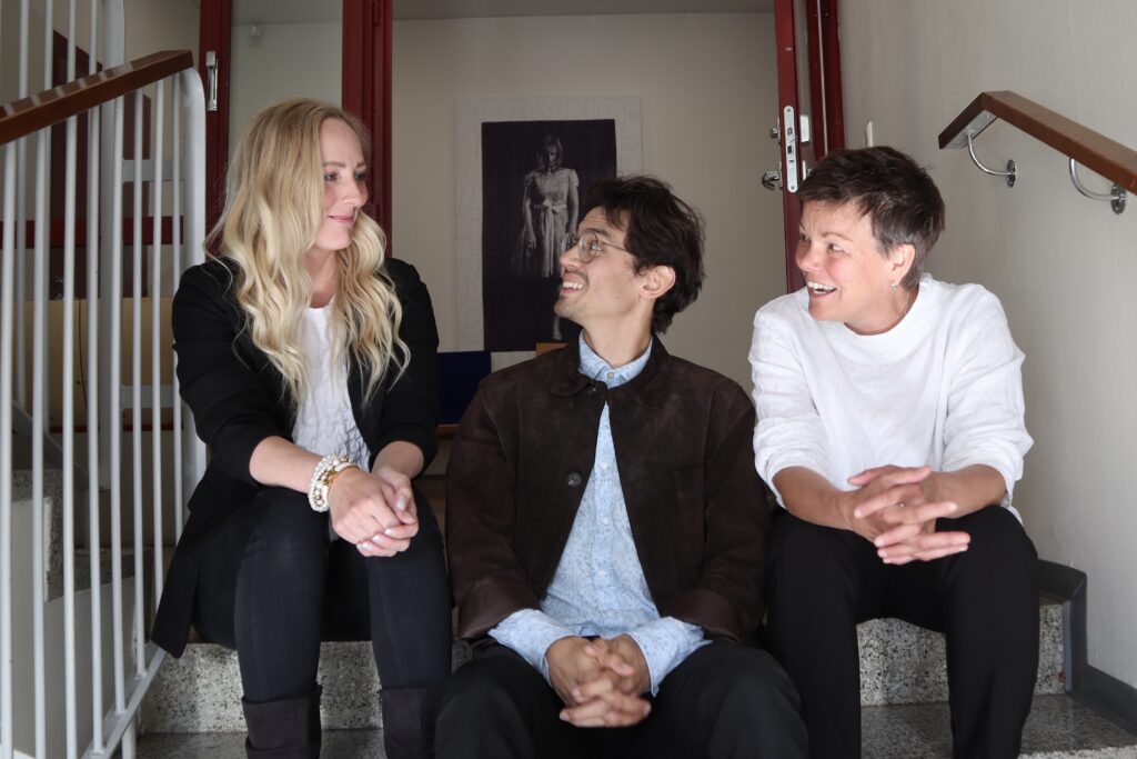 På bilden syns tre personer som sitter i en trappa inomhus. Längst till vänster finns en kvinna med långt ljus hår. Hon är klädd i mörk kavaj och mörka byxoir. I mitten sittern en unga man med mörk kavaj, mörkia byxor, ljusblå skjorta och glasögon. Han har mörkt lockigt hår. Han och kvinnan intill tittar på kvinnan till vänster. Längst till höger finns en kvinna med kort mörkt hår, vit blus och svarta byxor.