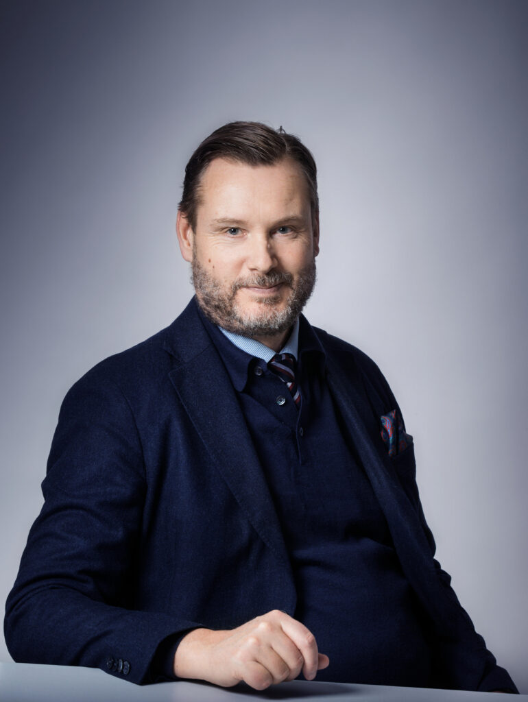 På bilden syns en man i medelåldern med kortklippt sakägg och brunt hår. Han är klädd i blå kostym, med en blå slkjorta. Han sitter vid ett bord med ena handen framför sig. Bakgrunden är gråblå i olika toner.