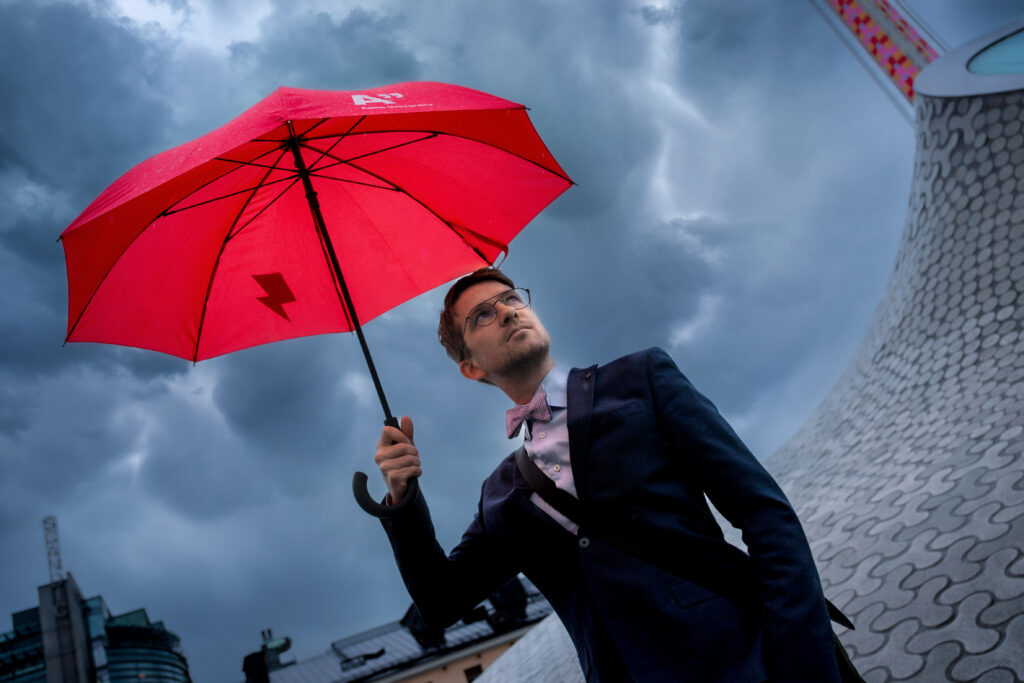 Man iklädd kavaj och fluga står med rött paraply, i bakgrunden syns en mörkgrå himmel.