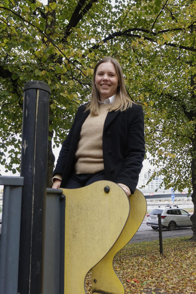 På bilden syns en kvinna, Sofia Jusslin som står högst uppe på en lekparksställning och tittar skrattande in i kameran. Hon har mellanblont långt hår och ör klädd i en mörk kappa och en ljusgul tröja. I bakgrunden ser man träd som skifter i grönt och gult.