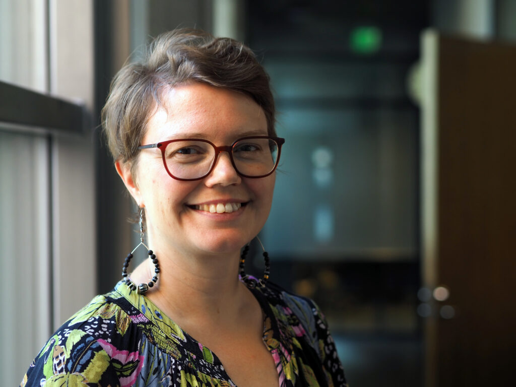 På bilden syns en relativt ung kvinna i kort mörkt hår och med bruna glasögon. Hon har stora örhängen, en mönstrad blus med svart botten och granns färger och hon ser glad ut. I bakgrunden syns kontorslokaler och fönster. Hon forskar i antirasism i skolan.