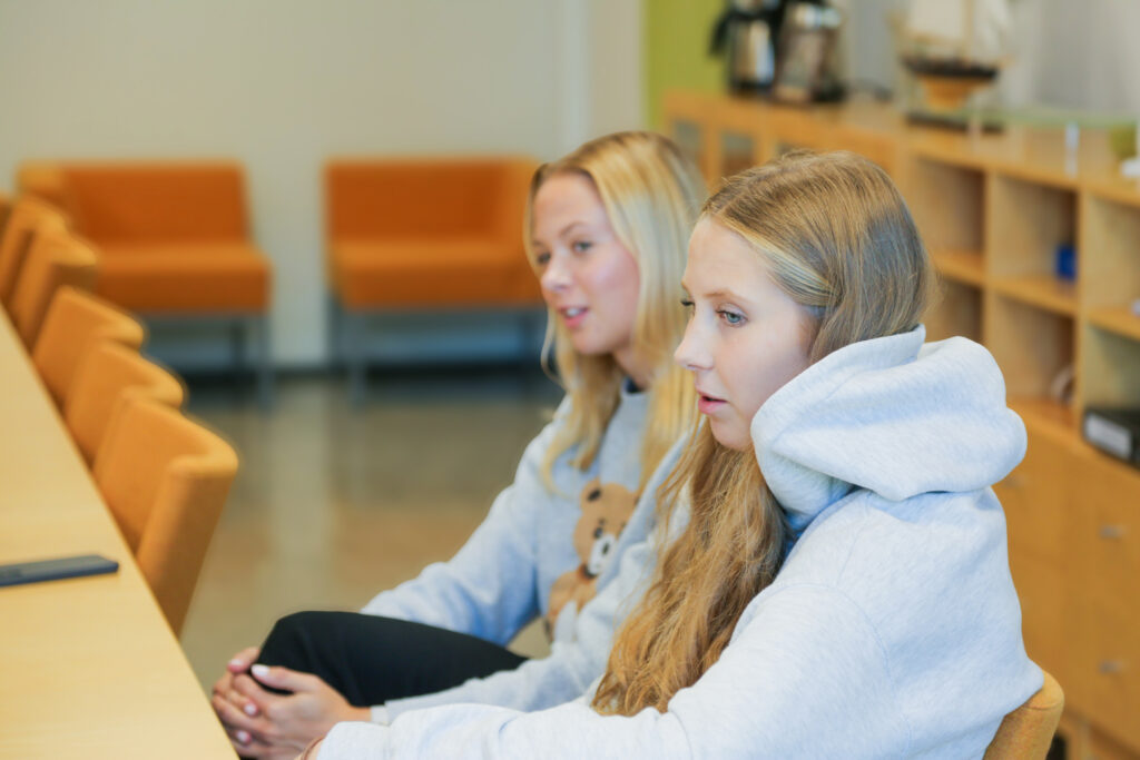 På bilden syns två blonda flickor som sitter och pratar vända lite åt sidan från kameran. De är båda klädda i gråa mjukiströjor. Bakgrunden är ett kontorslandskap med beiga väggar och golv och orangea stolar.