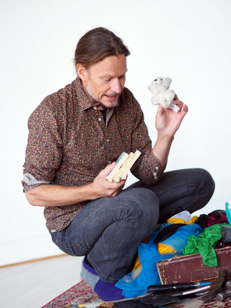 På bilden syns en medelålders man med långt brunt hår i knut. Han har ett kort skägg som skiftar i vitt. Han är klädd i brun skjorta med små röda blommor på och har mörkblåa jeans. Han sitter nedhukad framför en gammal brun resväska i läder som är proppfull av grejer. I en handen har han tre pocketböcker och i den andra en liten vit nallebjörn.