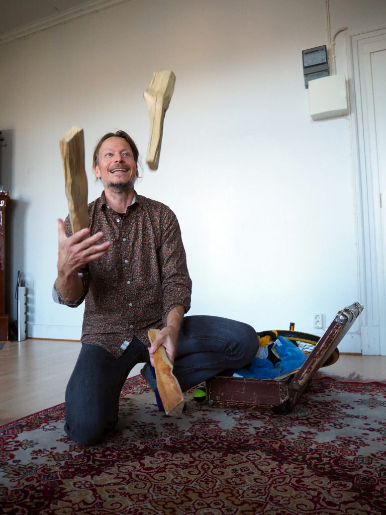 På bilden syns en medelålders man, Vesa Purokuru, som är kroppslig coach. Han har blåa ögon, långt brunt hår samlat i en knut i nacken och kort skägg som skiftar i vitt. Han är klädd i en brun skjorta med små röda blommor på och i mörkblåa jeans. Han står på huk bredvid en gammal resväska i brunt läder och han jonglerar med vedklabbar. På golvet syns en orientalisk matta i vitt, vonrött och ljusbrunt och i bakgrunden en vit vägg.