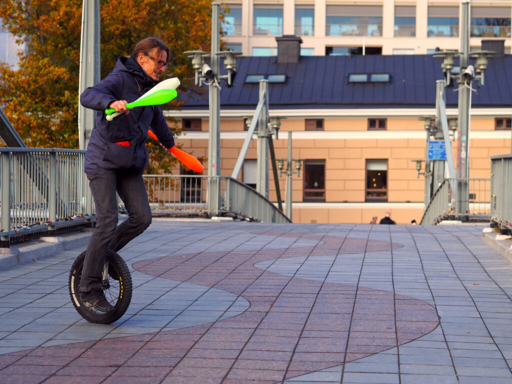 På bilden syns en medelålders man vid namn Vesa Puronkuru. Han är coach och talar för det kroppsliga lärandet. Han är klädd i en mörkblå jacka och har mörkblåa jeans. Han har glasögon, kort skägg som skiftar i vitt och långt brunt hår som är samlat i en knut i nacken. Han cyklar på en enhjuling med tjocka däck och i händerna har han tre käglor, en vit, en grön och en orange,