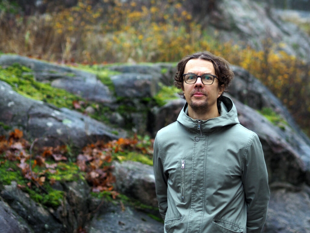 På bilden syns en man, Antti Kivijärvi, med lockigt brunt hår och mörka glasögon. Han har riktigt kort skägg och är klädd i en ljusgrön jacka. Han står framför en vy av klippor och växtlighet i höstfärger.