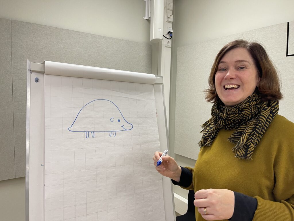 På bilden syns en medelålders kvinna med brunt hår. Hon tittar rakt in i kameran och ser glad ut. HOn är klädd i en gulgrön tröja och en scarf i samma färg och svart. Hon står vid ett högt block där hon ritat något som ska bli en igelkott.