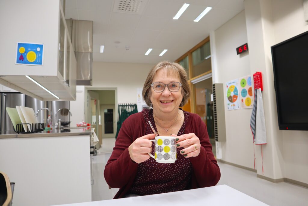 Kvinna med glasögon sitter leende med en kopp kakao i handen.