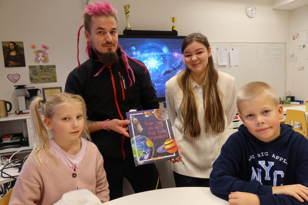 På bilden syns fyra personer som befinner sig i ett klassrum med ljusa väggar och ljusa möbler. I framre raden sitter två barn, En flicka klädd i rosa tröja och blus till vänster. Hon är blond och har håret uppsatt i två råttsvansar. Till höger sitter en blond pojke som är klädd i en mörkblå huvtröja. I bakre raden står till höger en man med rosa hår och brunt skägg. Han håller i en bok där det står Kan jag se mitt hus från rymden? Han är klädd i svarta kläder. Till höger står en kvinna med långt ljusbrunt hår. Hon är klädd i naturvit väst och blus och har mörka byxor.