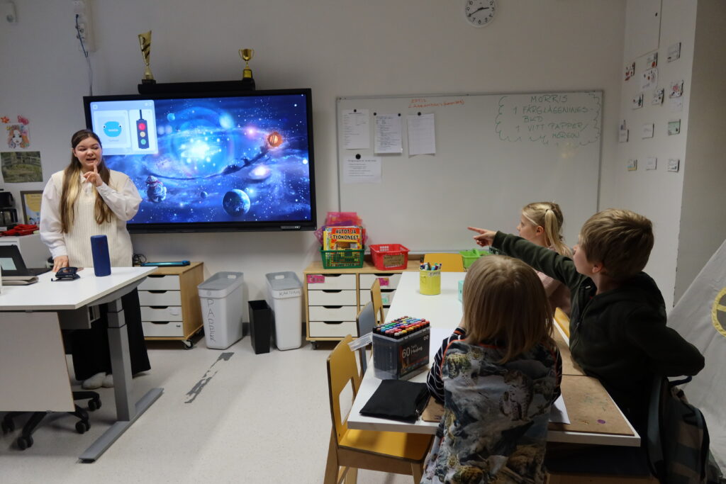 På bilden syns fyra personer som befinner sig i ett klassrum med ljusa väggar och vita bord. Till vänster står en kvinna som har långt brunt hår och är klädd i en naturvit tröja och blus och mörka byxor. Bakom hennen finns en stor skärm där man ser en tecknad bild av rymden med planeter och galaxer. Vi ett bord till höger i bilden sitter tre barn, en flicka med långt ljust hår i råttsvansar, och två pojkar med mellanbrunt hår som är klädda i märka tröjor.