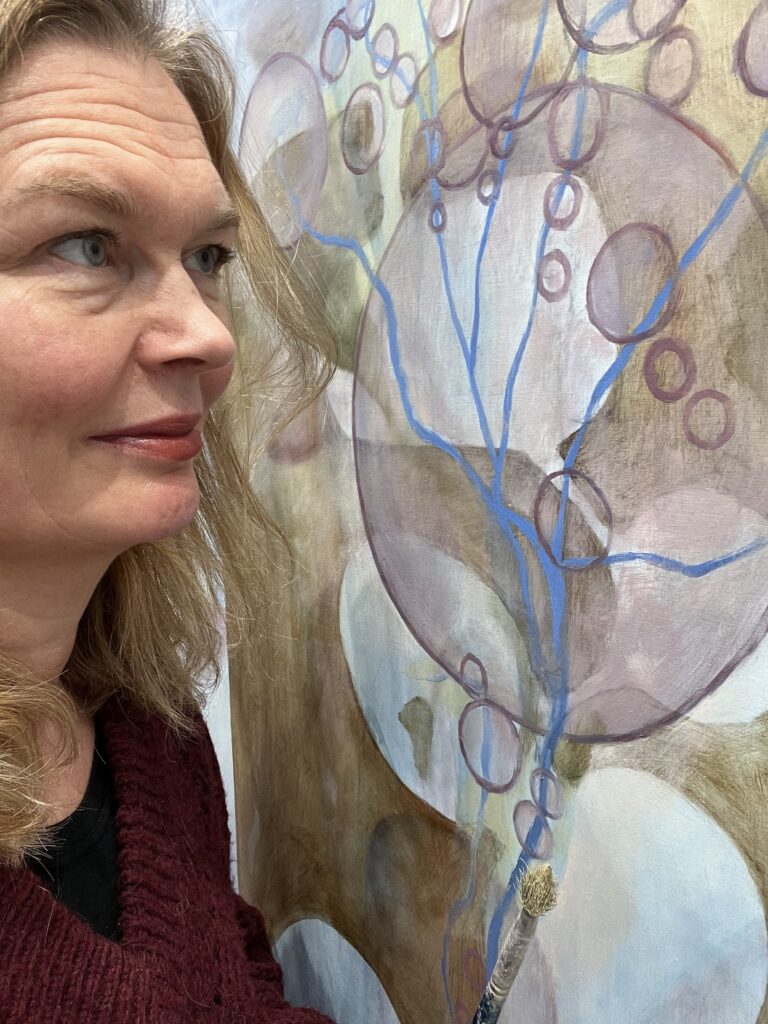 På bilden syns en medelålders kvinna med rödblont hår och gråa ögon. Bilden är tagen i närbild, halvt framifrån, så att man ser halv hennes ansikte. Hon tittar åt sidan och står framför en nonfigurativ tavla i beige, lila gult och blått.