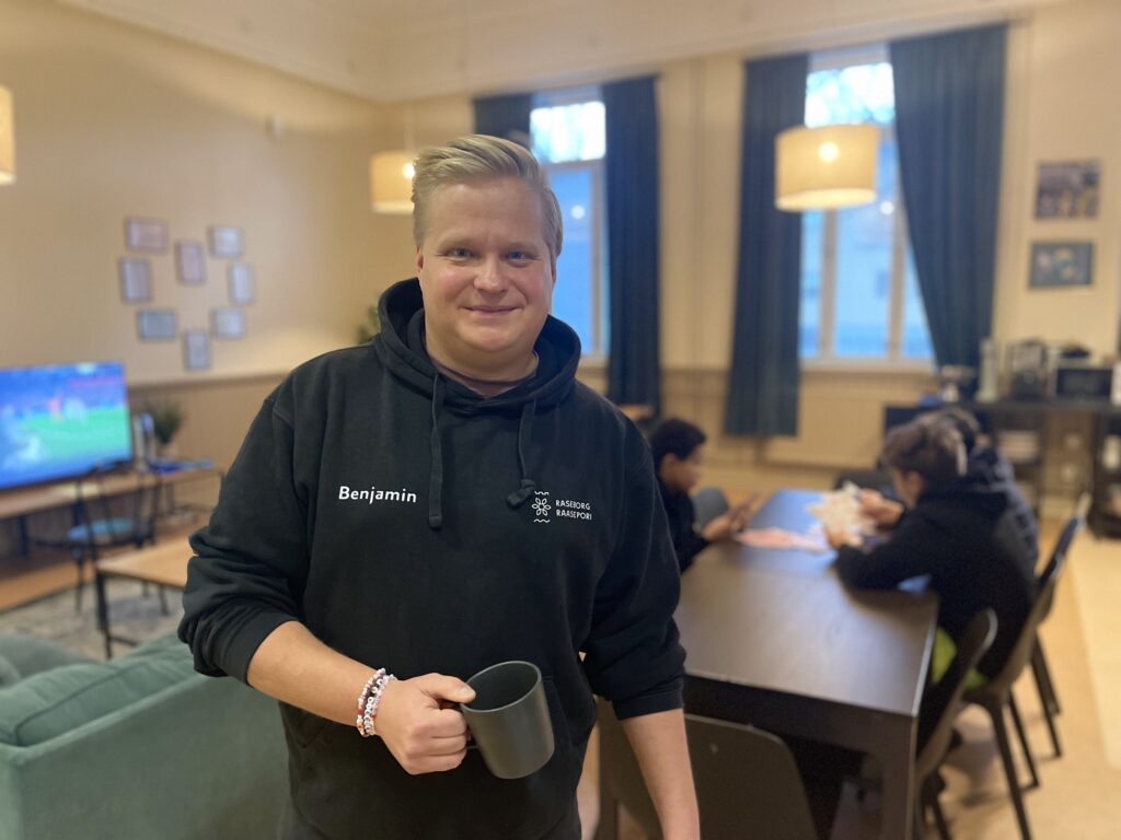 På bilden syns en blond man som tittar rakt in i kameran och ser glad ut. Han är klädd i en svart huvtröja, har en svart mugg i handen och armband i många färger på ena armen. I bakgrunden ser man en skärm, en grön soffa och ett rum med gula väggar och blåa gardiner. Man ser också ett bord där några pojkar spelar kort.