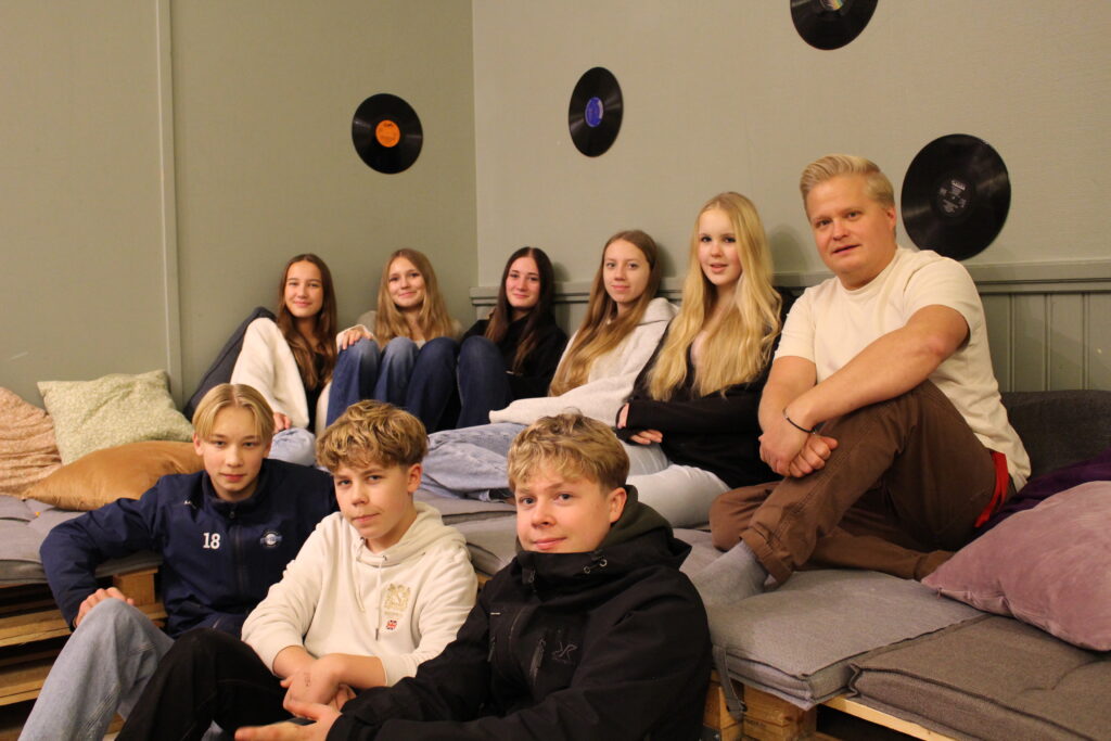 En grupp bild med fem flickor, en vuxen man och tre pojkar som sitter på och bredvid en soffa gjord av pallkragar. Väggen är ljusgrön och där finns lp-skicor uppklistrade på den.