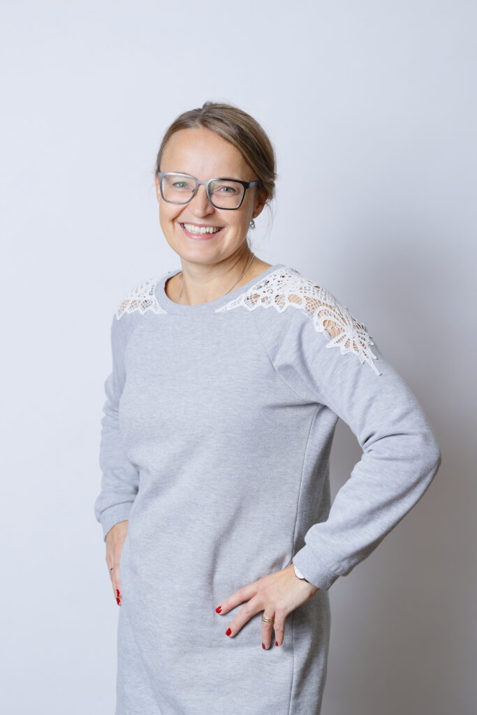 På bilden syns en kvinna med mörkblont hår som är draget bort från ansiktet. Hon har gråa metallbågade glasögon, ser rakt in i kameran och ler så man ser tänderna.
Hon är klädd i en grå rak klänning med vit spets på axlarna. Hon heter Mikaela Wiik och jobbar med frågor om arbetsvälbefinnande på Folkhälsan.