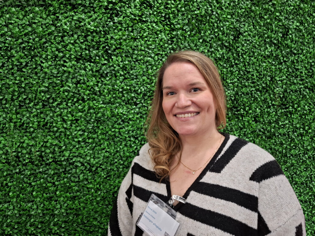 På bilden syns en kvinna vid namn Hann Viitala, universitetslektor i matematikens didaktik. Hon står framför en vägg av grönt plastgräs. Hon har långt ljust hår som är löst och draget fram över ena axeln. Hontittar rakt in i kameran och ser glad ut. Hon är klädd i en vit och svart tröja och i halsurringningen hänger en lapp med hennes namn på,