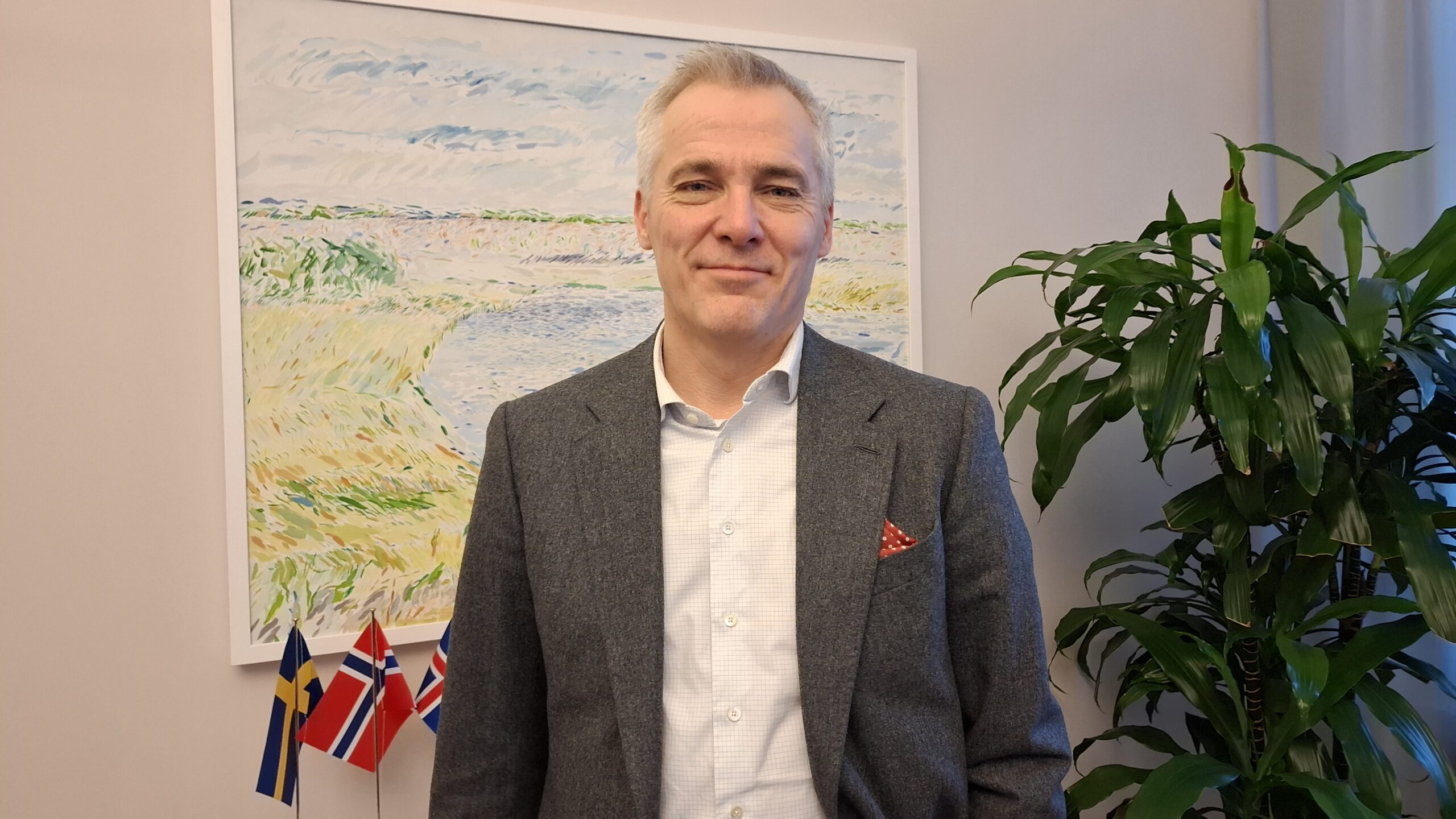 PÅ bilden syns en medelålders man, Anders Adlercreutz som är undervisningsminister i Finland. Han intervjuas om framtidsvisionen för den finländska grundskolan 20245. Han har grått och vitt kortklippt hår, ser rakt in i kameran och har en neutral min. Han är klädd i en grå kvaja med en röd och vitprickig näsduk i fickan och en ljus skjorta. I bakgrunden finns en mattrosa vägg, en tavla som föreställen en strand och några nordiska flaggor på ett bord. Man der också blad an en palmliknande växt.