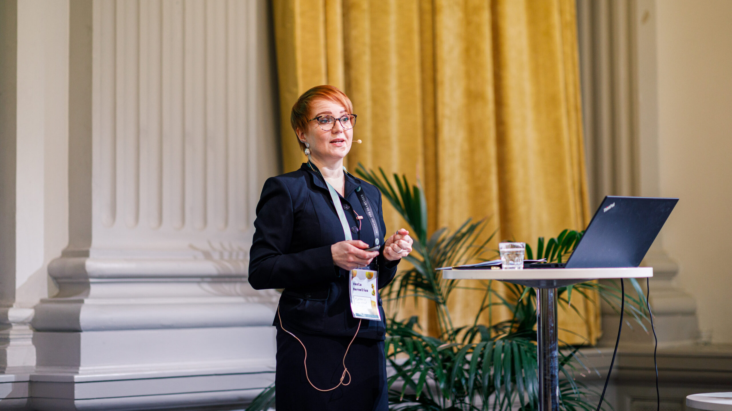 På bilden syns en ung kvinna vid namn Venla Bernelius, som är specialsakkunnig vid undervisnings- och kulturministeriet i Finland. Hon har kort rött hår, stora glasögon och silverörhängen. Hon har blicken fokuserad mot höger och står och pratar. Hon är klädd i mörkblå kavaj och kjol. Hon står vid ett talarpodium där det står en uppslagen laptop och ett glas vatten. I bakgrunden syns en vit utsirad pelare och ett gult draperi.