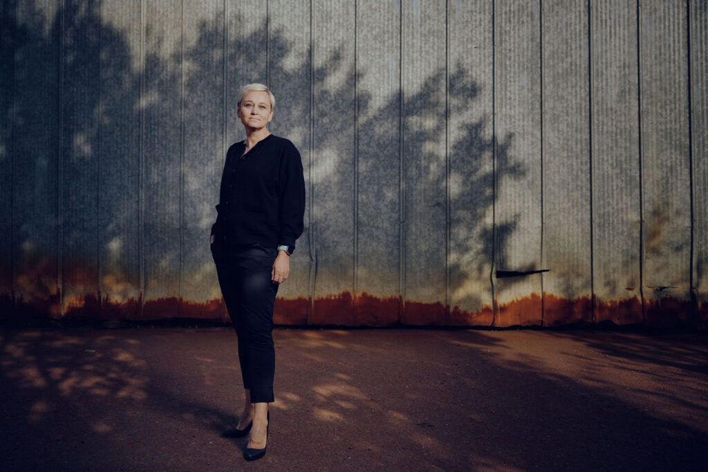 På bilden syns en kvinna i medelåldern i helfigur. Hon har kort ljust hår och är klädd i en mörkblå blus, mörkblåa byxor och mörkblå högklackade skor. Hon ser rakt i i kameran och ser allvarlig ut. Hon står framför en metall vägg i grått med en röd rand längst ner. Marken är röd asfalt. Mot muren syns skuggan av ett träd.
