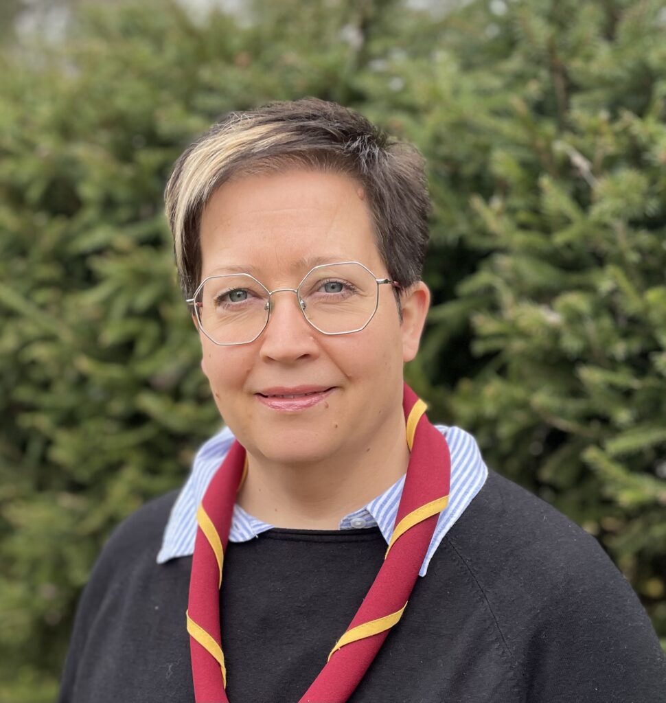 På bilden syns en kvinna i medelåldern, som heter Frida Nylund och jobba med scouting. Hon har kort mörkt hår med ljusa slingor i, kantinga glasögon med tunna ljusa metallbågar i grått. Hon ser rakt in i kameran och har en neutral min. Hon är klädd i en mörkgrå tröja med en ljusblå och-vitrandig skjortblus under. Runt halsen har hon en scouthalsduk i rött och gult. I bakgrunden ser man en grön häck.