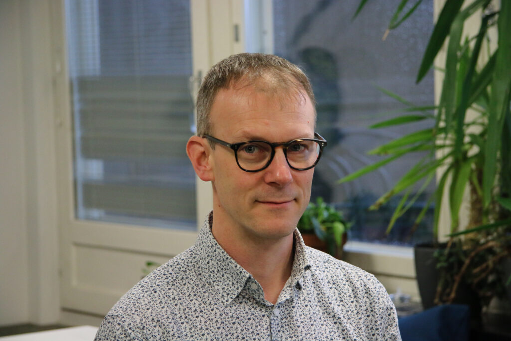 På bilden syns en man i trettioårsåldern, Kim Berts som är universitetslektor i matematikens didaktik. Han har tunt ljust hår, mörka glasögon och tittar lite klurigt in i kameran. Han är klädd i en skjortblus med vit botten och mörkblått mönster. Han är fotad inomhus, bakom honom ser man blurrat en balkongsdörr och ett par gröna krukväxter.
