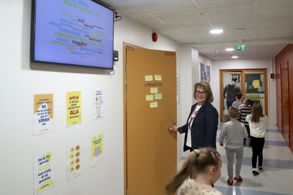 På bilden syns en korridor i en skola. Till höger finns en infotavla med ord i olika klara färger. Under den finns sex planscher i orange, gult och blått som är upphängda på en vit vägg. Bredvid planscherna finns en gulbrun dörr där det står Lärarrum. Den har gula och gröna postitlappar på. En äldre kvinna i blå kavaj och vit blus står och håller i dörren. Hon tittarmot kameran och ler. Bredvid henne i korridoren går en kö av barn efter en manlig lärare. Barnen ser ut att vara ungefär åtta åt gamla . Längt still höger ser man en rödmålad vägg med rödmålade dörrar.