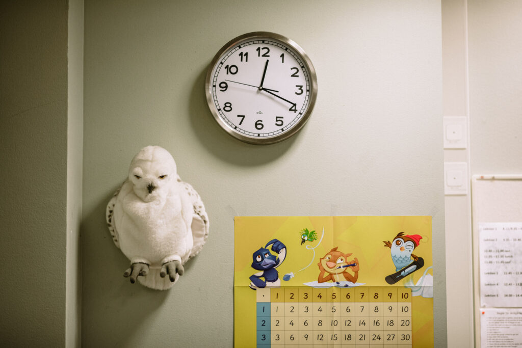 BIlden föreställer en vägg i ett klassrum. Överst syns en analog klocka med metallram, vit botten och svarta siffror. Klockan visar tjugo över tolv. Snett nere till vänster finns en mjukisuggla i vitt och beige och bredvid den till höger en multiplikationstabell på gul botten och med glada ritade djurfigurer som räknar matte och snowboardar. Bilden illustrerar en artikel som handlar om rektorns roll för välbefinnandet i skolan.