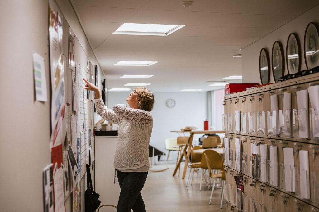 På bilden syns en lärare som står och tittar på ett schema på en vähh i ett lärarrum. Hon har pekfingret på schemat och ser fundersam ut. Man ser henne från sidan. Hon har ljus, kort lockigt hår, mörka glasögon, en vit tröja med spetsmönster och svarta byxor. Till höger i bilden syns en rad med skåp och iovanför dem svartvita porträtt. I bakgrunden blurrat syns bord, stolar och ett kök. Väggarna är ljusa i beige och gröna toner. Bilden hänför sig till en artikel som handlar om rektorns roll för välbefinnandet i skolan.