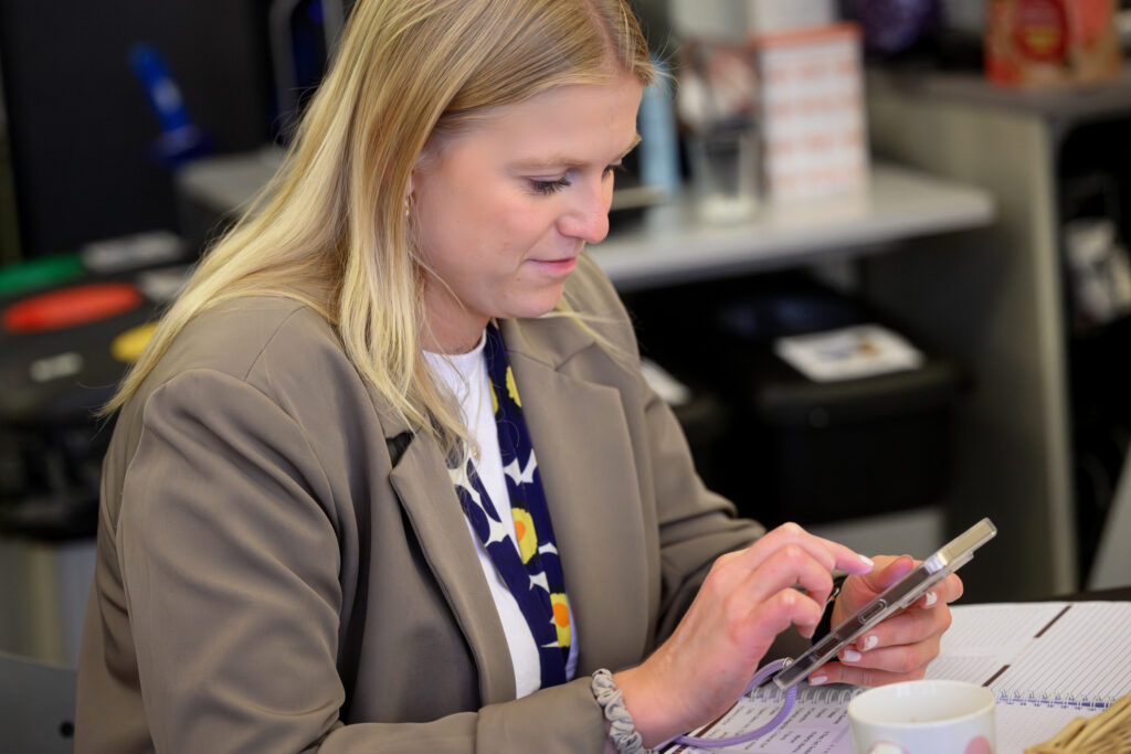 På bilden ser vi en kvinna som sitter och skriver på en mobiltelefon. Kvinnan har långt ljust hår som är löst. Hon tittar koncentrerat på sin telefon och ser nöjd ut. Hon är klädd i en beige kavah och har en vit t-shirt under. Runt halsen har hon ett nyckelband med Marimekkos Unikko-motiv i blått och gult. I bakgrunden ser man suddigt en miljö som ser ut som ett lärarrum.