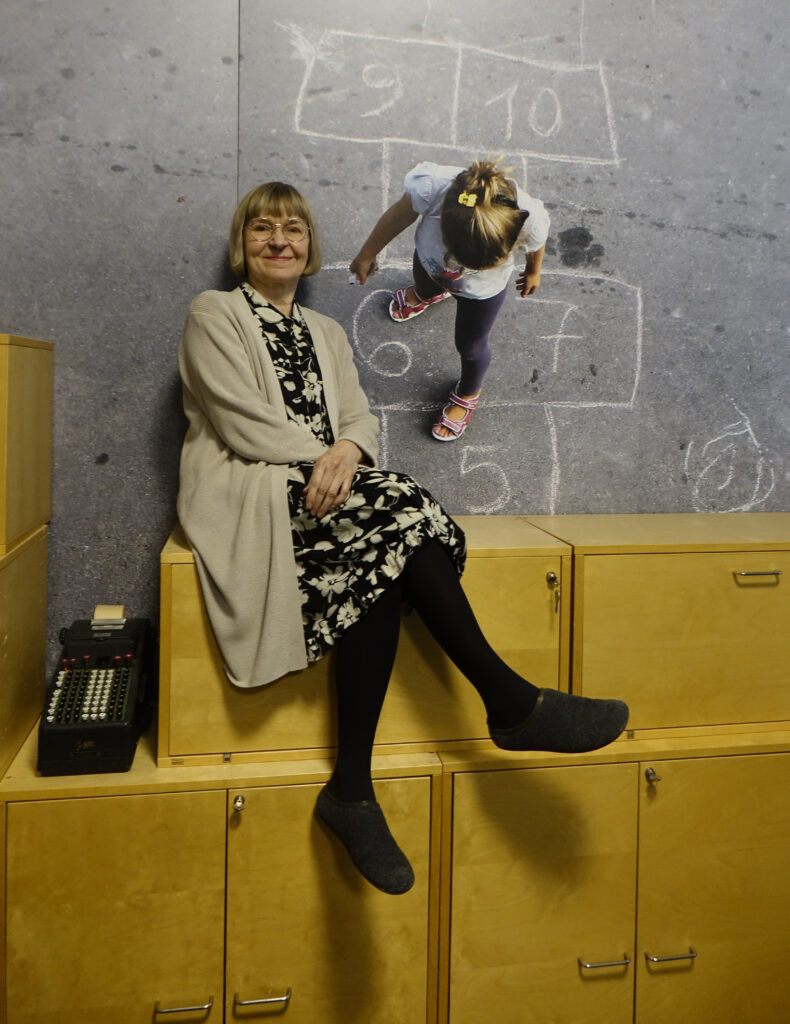 På bilden syns en äldre kvinna med pageklippt ljust och grått hår, tuglasögon med tunna metallbågar. Hon är klädd i en blommig klänning med gula blommor på svart botten och ovanpå det en beige tröja. HOn sitter på en hylla i ljust trä framför en stor väggbild av en liten blond flicka som hoppar ruta på en grå asfaltsyta. Kvinnan heter Tuija Metso och är sakkunnig på Finlands föräldraförbund. Hon talar om kommunikatione i Wilma mellan skola och hem. Hon tittar rakt in i kameran och ser glad ut.