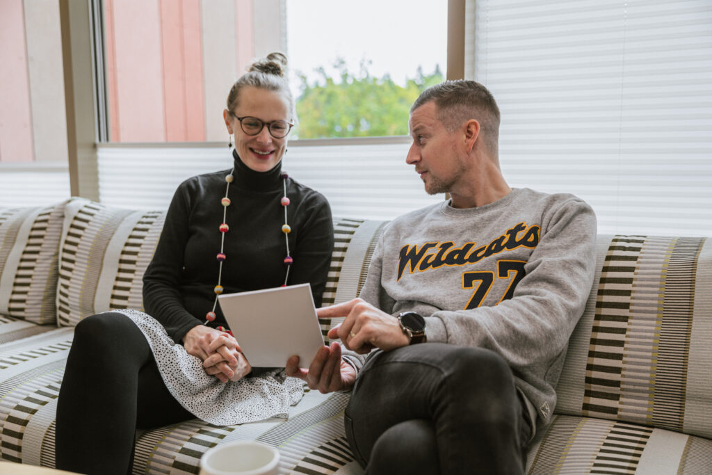 På bilden ser vi två personer som sitter på en randig soffa framför ett fönster med spjälgardiner. Kvinnan till vänster har en svart poloblus, vit kjol med svarta prickar och svarta strumpbyxor. Runt halsen har hon ett färggrant halsband med bollar på en lång tråd. Bredvid hennen sitter en man i lägre medelåldern som håller i ett häfte och pekar på det. Han tittar på kvinnan som tittar på häftet. Mannen är kortklippt och har mörkt hår. Han är klädd i en ljusgrå collegeblus med texten Wildcats på i svart och gult. Han har svarta jeans.