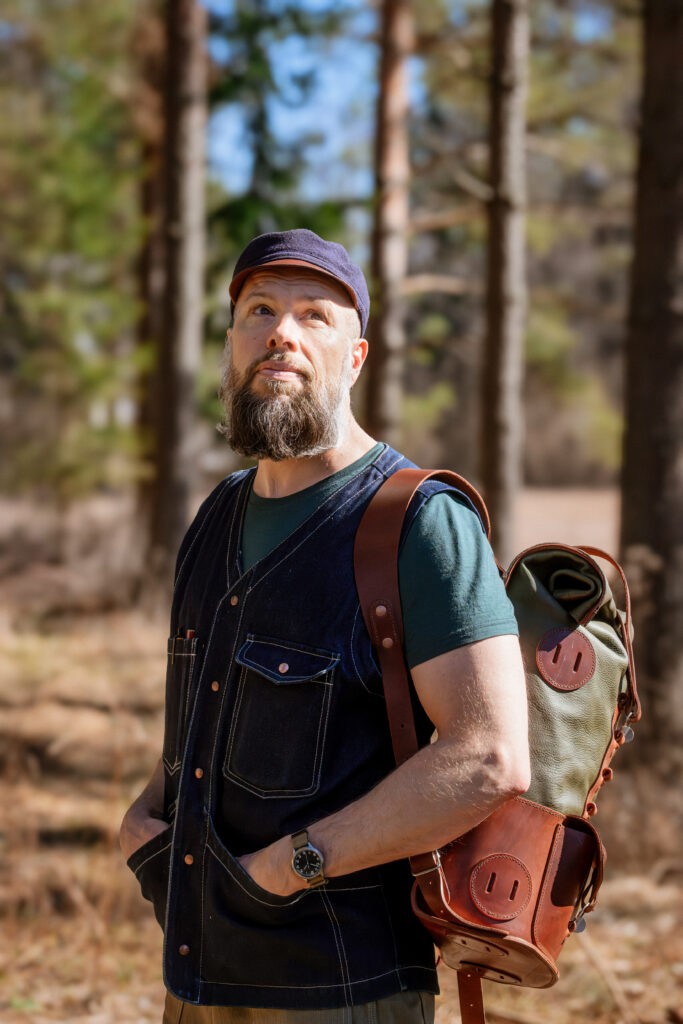 Man med skägg iklädd keps står i skogen med en läderryggsäck på ryggen.