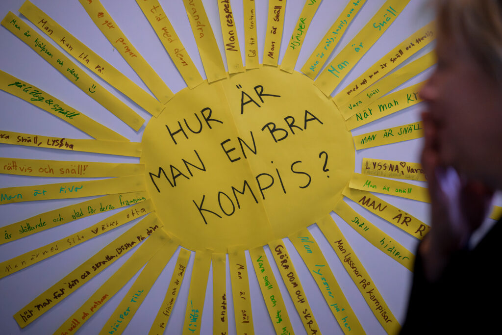 På bilden syns en sol utklippt i gult papper som är uppsatt på en vit vägg. Mitt i solen finns texten: Hur är man en bra kompis? och på strålarna har elever skrivit olika svar på frågan. Framför solen ser man en suddig figur av en elev.