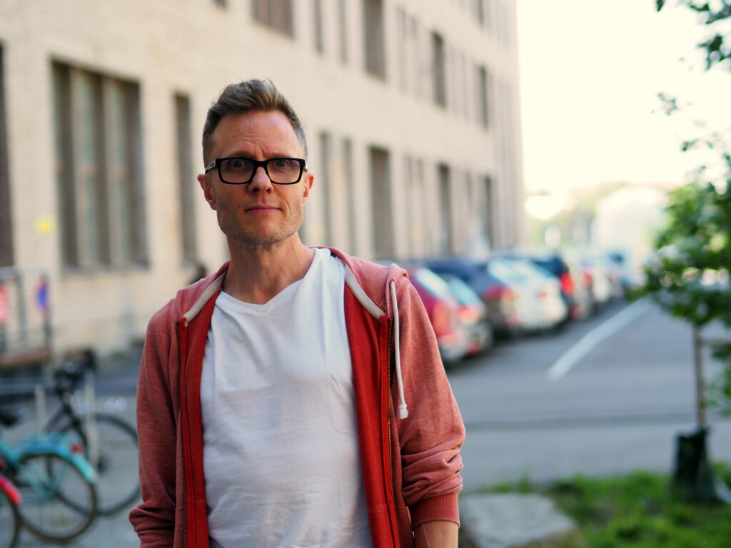 På bilden syns en medelålers man, Tom Pakkanen som talar om våld i skolan. Han har kort brunt hår som står uppåp mitt på huvudet. Han har glasögon med mörka tjocka skalmar och är klädd i en vit t-shirt och en huvtröja i rösa och rött. Han är fotograferad utomhus och i bakgrunden ser man suddigt ett ljust hus och parkerade bilar och cyklar.